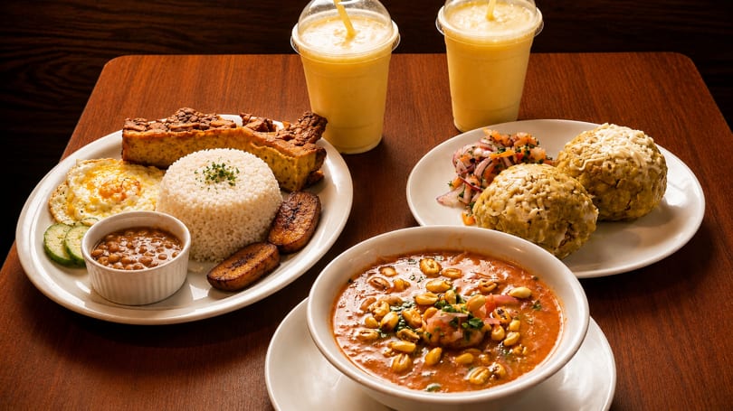A table laden with various dishes, including rice, beans, plantains, soup, and two yellow drinks.