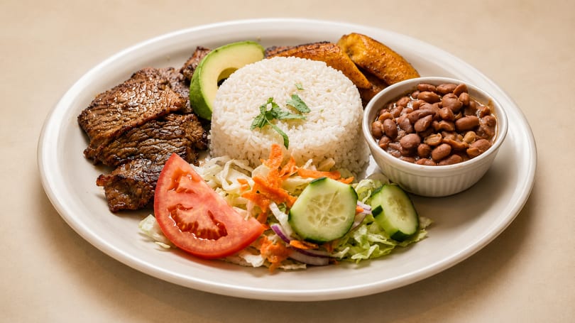 Grilled steak, rice, beans, avocado, plantains, and salad on a white plate.