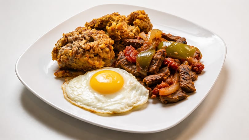 A white plate with a fried egg, beef and peppers, and a side of mofongo.
